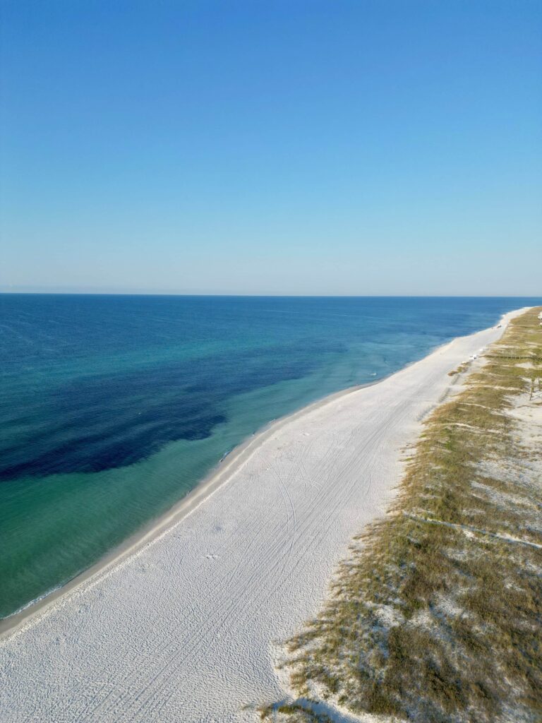 A drone photo above Pensacola Beach with white sand, blue sky, and emerald ocean water