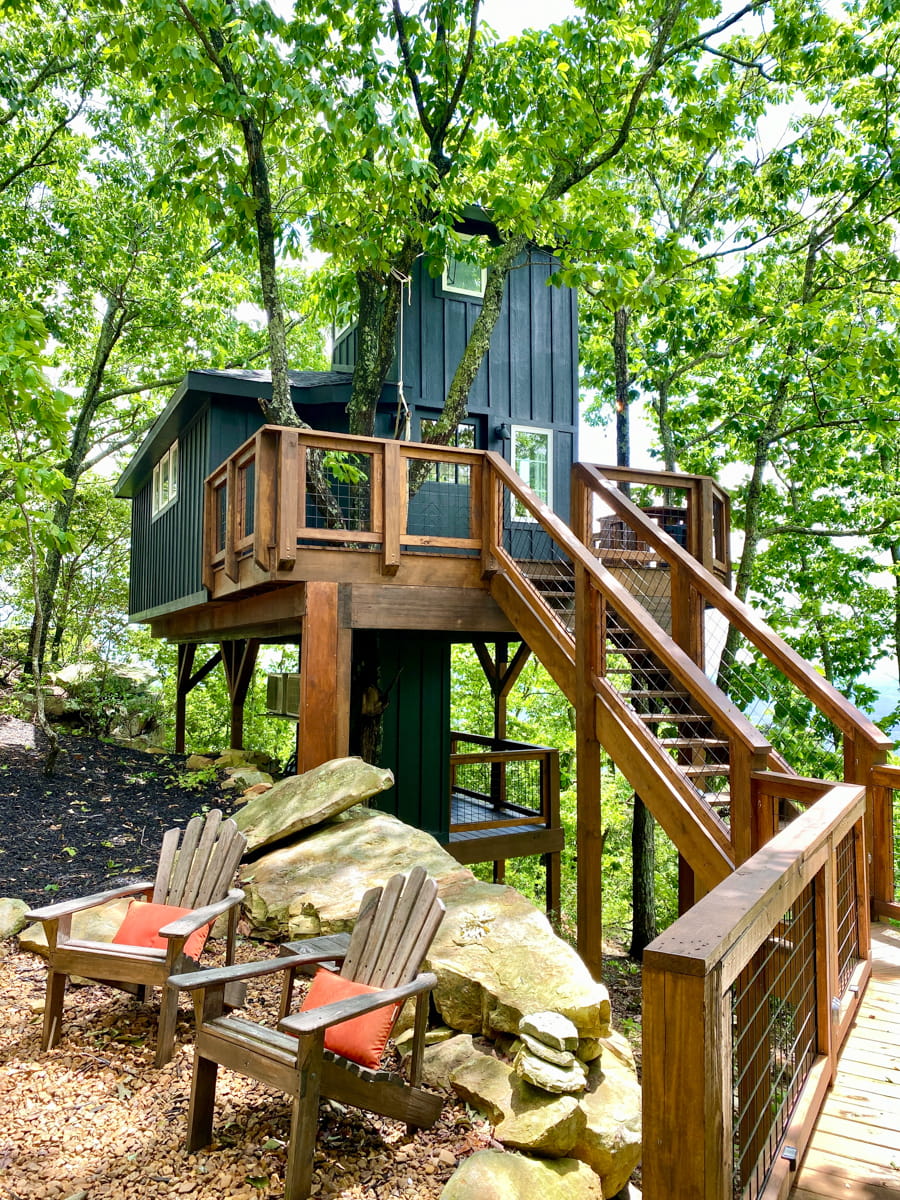A wooden staircase leading up to a black treehouse structure with woods around it and two Adirondack chairs beside the staircase
