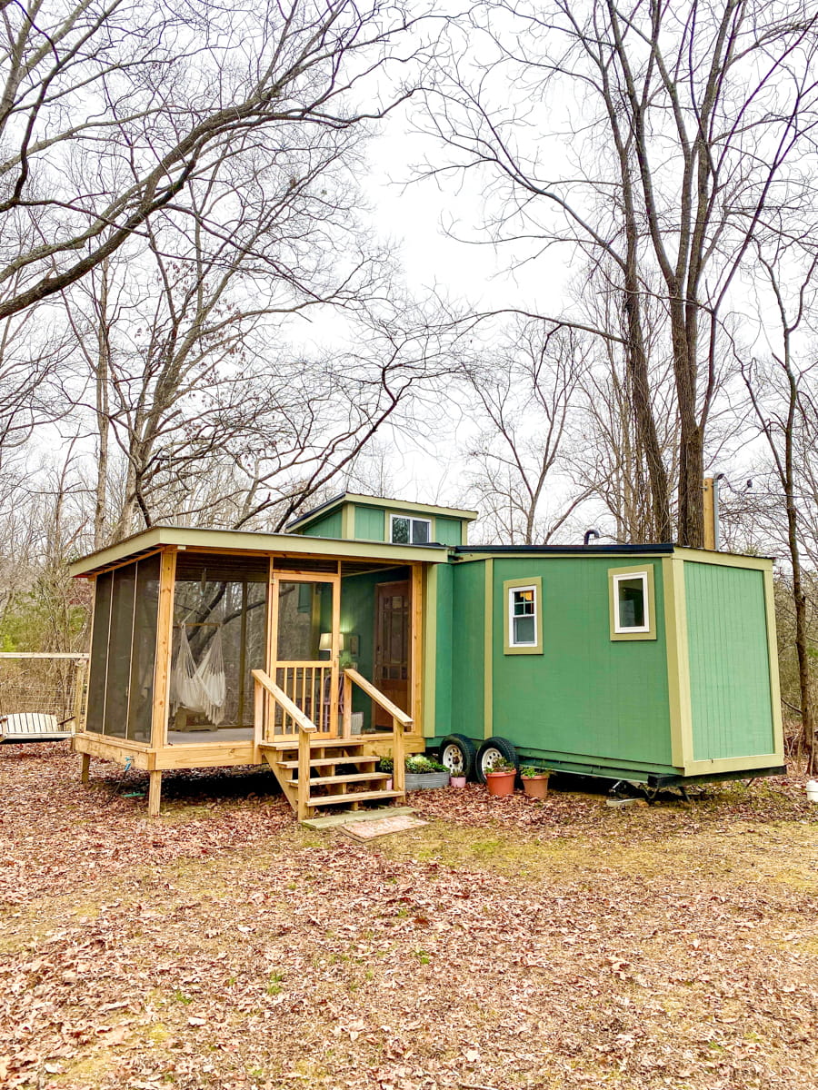 A green tiny house with a screened-in porch and winter forest behind it