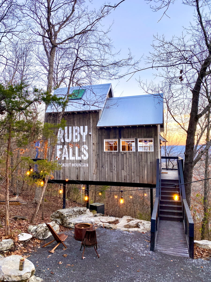 A wooden black staircase leads up to a treehouse with the name RUBY FALLS on the siding with a winter forest and sunset view behind it