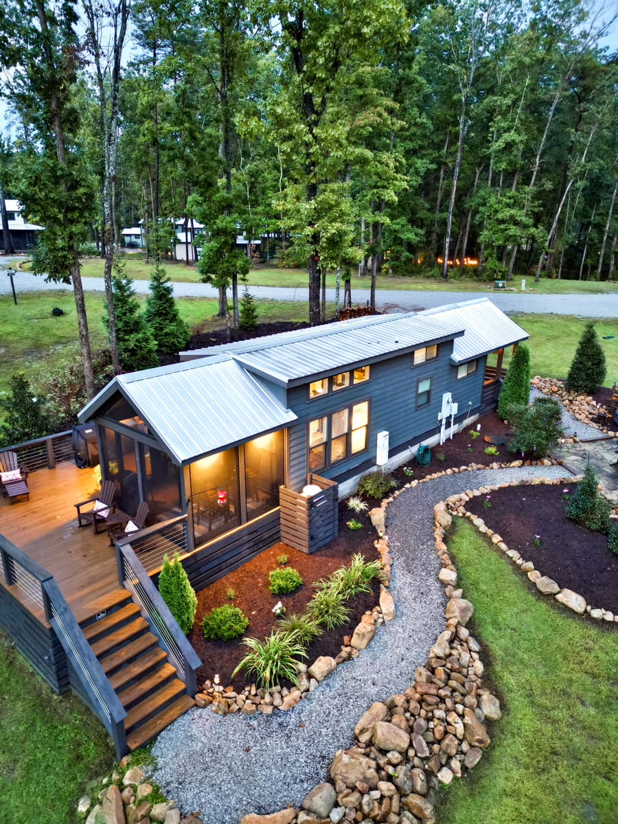 A retangular gray tiny home with outdoor lights at dusk and a porch deck with pathway in the front
