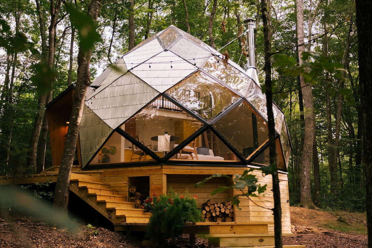 The exterior of the Airbnb with glass windows, string lights, a pointed roof, and wood stacked underneath the structure with a staircase leading down to the hot tub